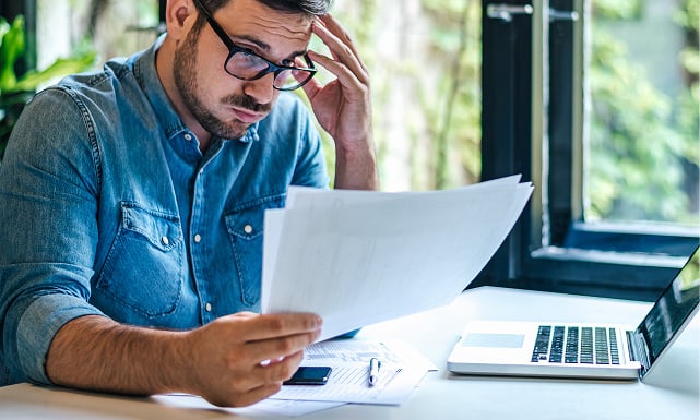 A man sitting at a desk with a confused expression looking at printed documents, and there's a laptop, pen, cellphone, and more documents on the desk.